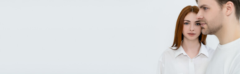 Red haired woman looking at camera near blurred boyfriend isolated on white, banner.