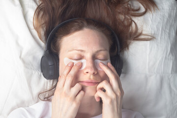 A Caucasian woman with gel patches on her eyelids to care for the skin around her eyes lies and listens to music with headphones