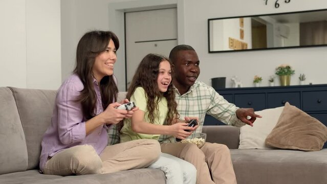 Multi ethnic family playing video game at home sitting on sofa. The daughter wins over the mother and rejoices. Game On, Family Meeting, Multi Ethnic Family, Different Generations.