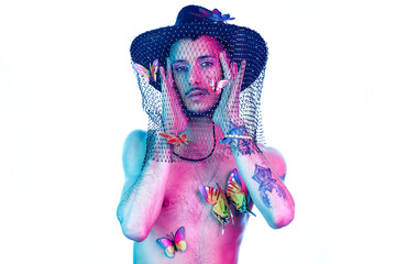 Young model with tattoos, wearing a hat with a mesh, hanging butterflies as decorative accessories and makeup. Posing with white background, magenta and blue light. Showing her feelings.