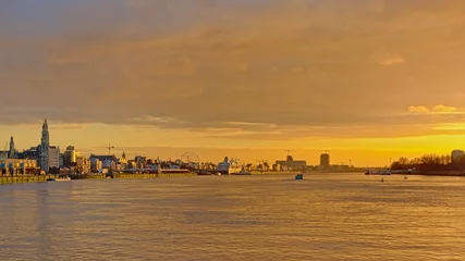 Fotobehang De skyline van Antwerpen met reuzenrad, wolkenkrabbers en cruiseschip, uitzicht vanaf de overkant van de Schelde in warm oranje licht na zonsondergang © Kristof Lauwers