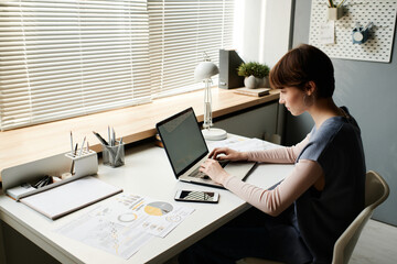 Concentrated young businesswoman sitting at desk and using laptop while communicating with client...