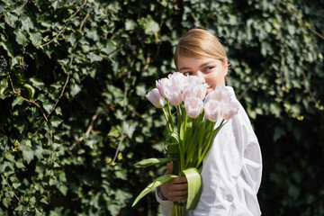 happy young woman obscuring face with white tulips near green ivy.