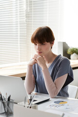 Concentrated young female manager in casual vest sitting at table with graphs and examining report on laptop