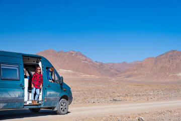Woman in a red checkered sweater with her son leaning out the window and door of a campervan with a dusty road ahead