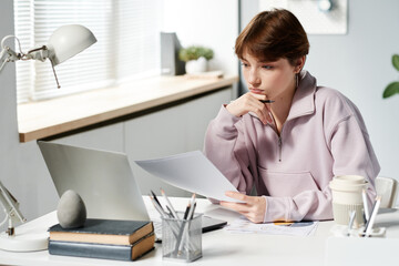 Serious Caucasian girl in hoodie sitting at desk and looking at laptop screen while analyzing budget in office