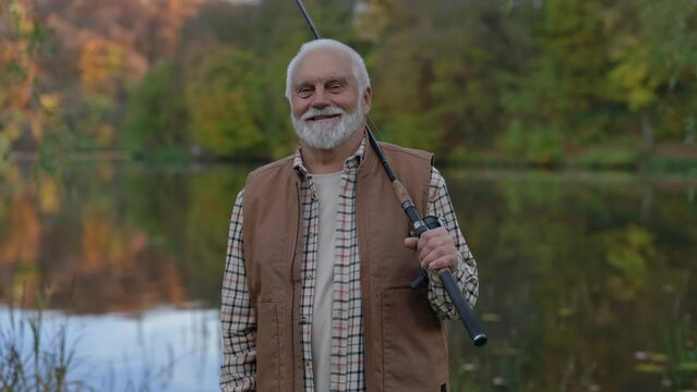 Portrait of grey haired professional fisherman holding spinning rod while looking at the camera near lake. Caucasian aged male enjoying happy retirement on fresh air.