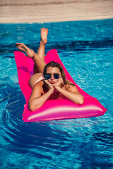 Sexy woman in a swimsuit lies on a pink inflatable mattress in the pool. Relax by the pool on a hot summer sunny day. Vacation concept