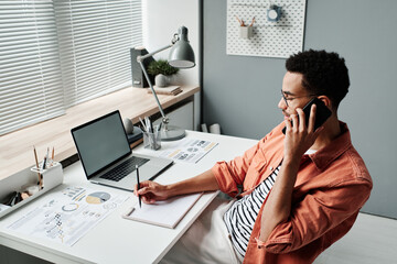 Serious young black businessman in stylish shirt talking by phone while scheduling meeting in office