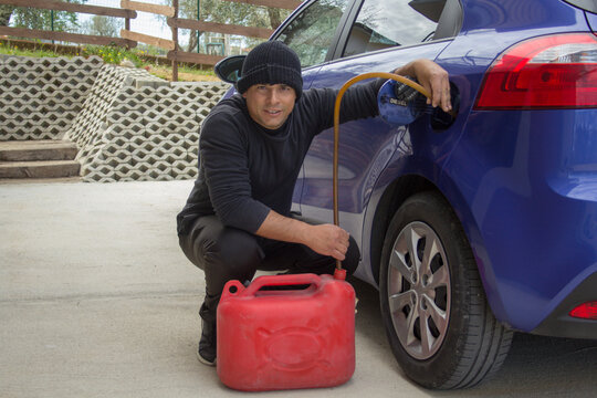Thief man dressed in black sucking fuel from a parked car. Reference to expensive gasoline