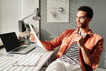 Focused young African American manager with stubble sitting at desk against blinds and examining...