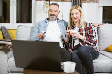 Beautiful married couple of two mature people in casual wear sitting together on couch and watching film on modern laptop. Happy caucasian family spending free time together at home.