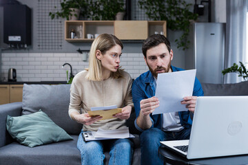 Young family, husband and wife, reviewing their bills, loan and mortgage agreements, sitting on the couch at home, working with documents on a laptop, embarrassed, upset