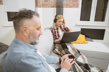 Attractive mature woman sitting on comfy couch in headphones and typing on laptop while her handsome husband using smartphone. Family at home with modern technology.
