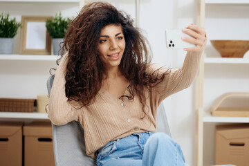 Cheerful awesome tanned curly Latin lady taking selfie with trendy smartphone sit on chair in home modern interior look aside. Copy space Mockup Banner. Concept time to love yourself