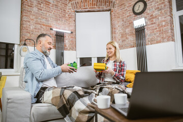 Positive mature family making surprise to each other with presenting gift boxes during their anniversary at home. Handsome man and charming woman sitting together on couch and smiling to each other.