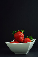 Red, juicy, appetizing strawberries in a white porcelain bowl. Vertical shot, black background with copy space.