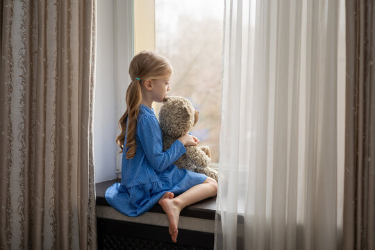 A little girl with long blond hair sits on the windowsill with a toy and looks out the window into the street. This is a sad mood. Illness or isolation.