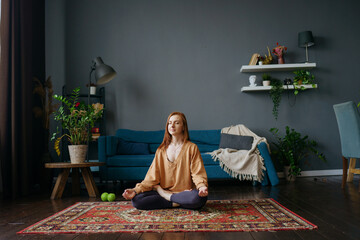A woman with red hair sits in a lotus position on the floor and meditates in her living room from afar