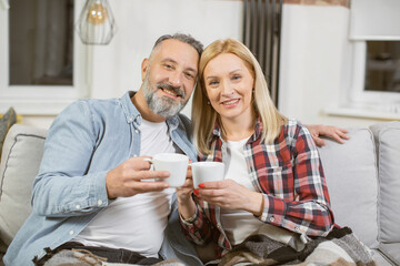 Middle aged couple in casual wear sitting on cozy sofa with cups of drink in hands. Beautiful caucasian family resting together at home during leisure time.