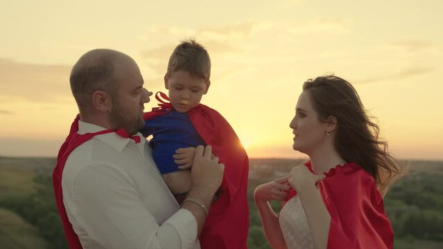 Father and son, mother, play superheroes at sunset. Happy family in superhero red capes are playing in park in sun. Friendly family concept. Happy childhood. Mom dad kid dream together, family team