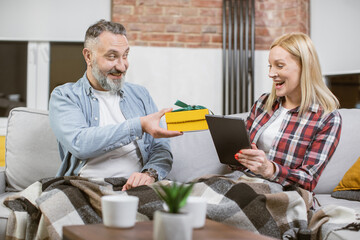 Caucasian caring man presenting gift box to his charming woman that sitting on couch with tablet in hands. Attention, surprise and family concept.