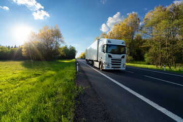 Fototapeta na wymiar White truck driving on the asphalt road between the forest and the meadow in springtime landscape at sunset