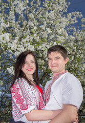 Young loving couple man and woman in white embroidered shirts near a flowering tree in a spring garden.
