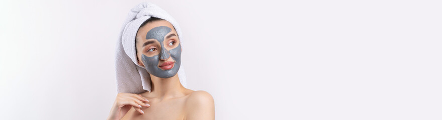 A woman with a gray clay cosmetic mask in a white towel on her head on a white background.