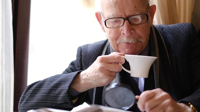Senior man holding a cup of coffee sitting at a table in a cafe reading a newspaper.