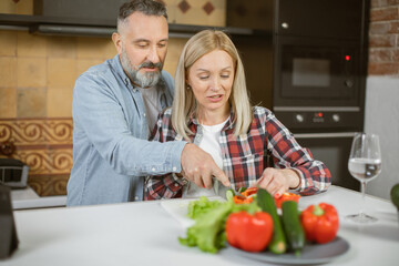Happy mature couple in love chopping fresh vegetables on kitchen and using digital tablet. Caucasian family in casual wear enjoying common time at cozy home.