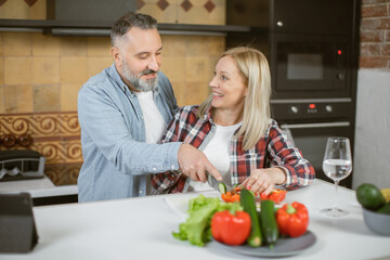 Happy mature couple in love chopping fresh vegetables on kitchen and using digital tablet. Caucasian family in casual wear enjoying common time at cozy home.