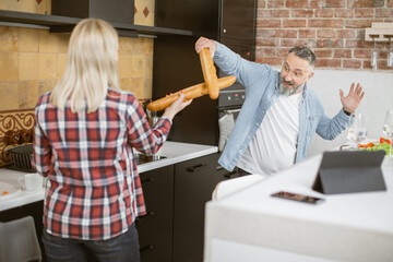 Joyful mature man and woman fighting with baguettes like as swords during cooking process on...