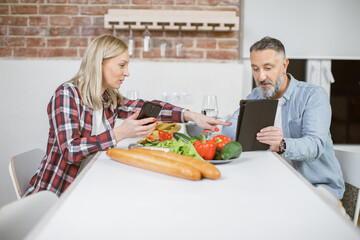 Middle aged man and woman sitting together at kitchen table and using modern smartphone and digital table. Concept of family, leisure time and technology.