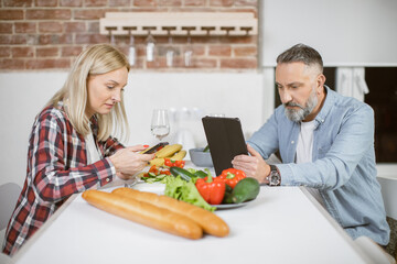 Middle aged man and woman sitting together at kitchen table and using modern smartphone and digital table. Concept of family, leisure time and technology.
