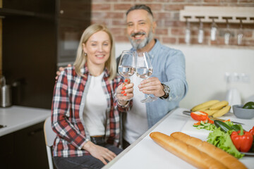 Happy mature couple in casual attire making toast with glasses of water for their healthy lifestyles. Portrait of caucasian family sitting at kitchen table with fresh organic vegetables their.