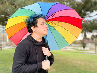 A smiling young woman poses with an open rainbow umbrella in a park. Dressed in black casual clothes and with blue hair. Diversity and respect concept.