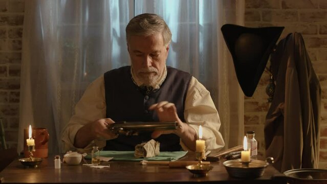 A mature bearded man from the 1700s polishing his flintlock pistol as part of maintenance on the firearm.