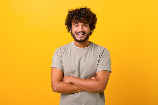 Indian Youngster Looking At Camera Standing With Arms Crossed Isolated On Yellow Background. Eastern Male Student With Curly Hair In Casual T-shirt With Folded Hands