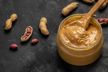 Peanut butter or paste in an open jar and peanuts in the peel scattered on a dark background, banner, menu, recipe place for text, top view