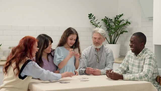 Multi ethnic family of different age generations play cards at home sitting at the table. The child wins. Game On, Family Meeting, Multi Ethnic Family, Different Generations.