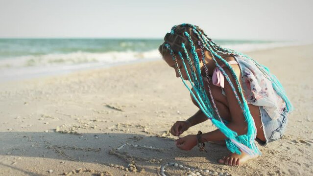 A girl with african braids in a summer costume plays on the beach with shells near the sea with waves under sunny sunset