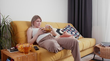 A pregnant young woman is sitting on the couch, eating popcorn and crying while watching TV. A young woman is pregnant.