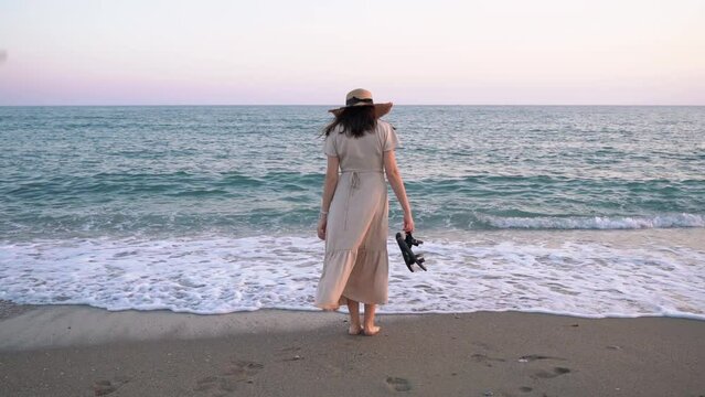 Rear view caucasian slim lady standing in front of Mediterranean sea coast. Happy woman in summer dress holding sandals and enjoying the sunset at the beach.