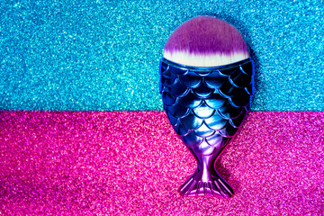 Brush to liquid base. Liquid foundation brush. Makeup brush to apply liquid foundation on the skin. Blue and pink fish-shaped brush, on a colorful glitter background.