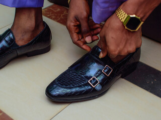 An african nigerian groom or man with a watch on his wrist, wearing his wedding shoe as he prepare...