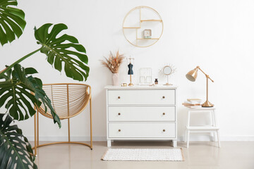 Chest of drawers with stylish jewelry, houseplant, armchair, stepladder stool with books and lamp near light wall in room