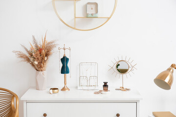 Stands with stylish jewelry, vase with flowers, mirror and perfume on white chest of drawers near light wall