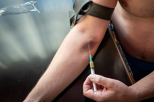 Addict injecting heroin dose to his hand with syringe. Man drugs addicted injecting heroine in his arm at the table with other drug on black background in dark