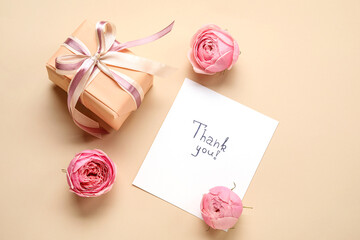 Sheet of paper with text THANK YOU, gift box and flowers on beige background